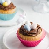 A yellow cake cupcake topped with chocolate buttercream frosting, three Cadbury mini eggs, and an Easter bunny party pick, with another cupcake in the background